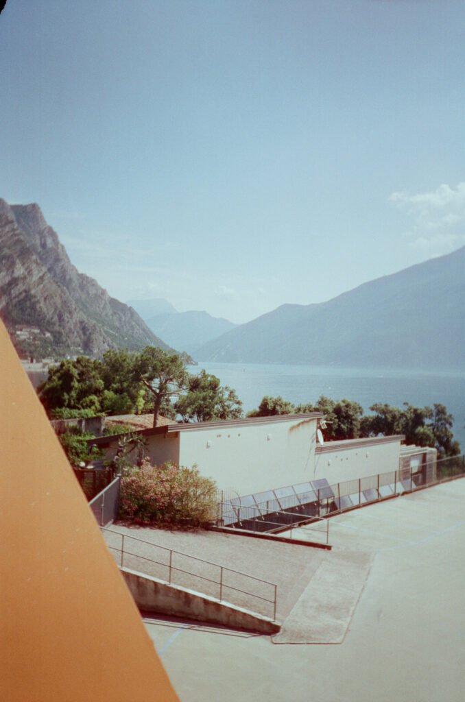 Elevated daytime view over Lake Garda from a lakeside terrace, with a flat-roofed building and solar panels below, olive trees and an oleander in bloom, and steep hazy mountains rising from the blue water.