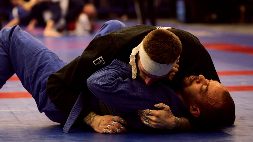 Two grapplers in gis on the competition mat, the top player in a black gi controlling the head of an opponent in a blue gi.