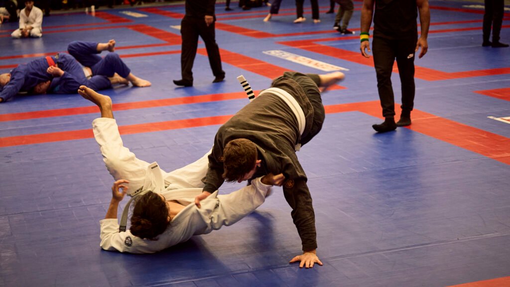A jiu-jitsu match in progress, a competitor in a dark gi sweeping an opponent in a white gi to the mat as others train behind.