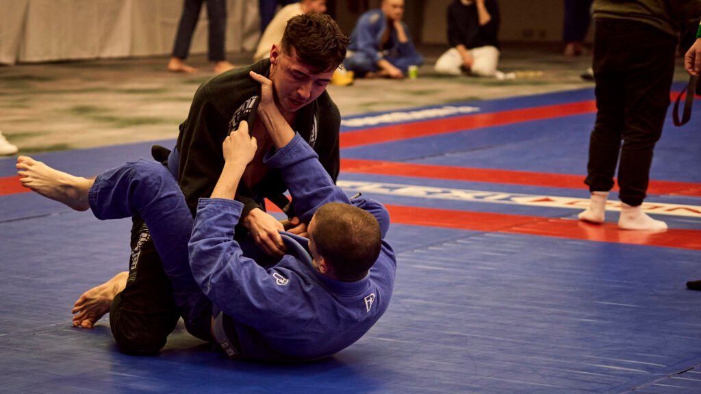 Two competitors in gis grappling, the top player gripping the collar of an opponent playing guard on the blue mat.
