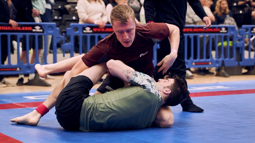 A no-gi grappling match, one competitor in a maroon shirt controlling an opponent on his back on the blue mat at a tournament.