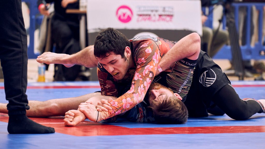 A no-gi competitor in a floral rashguard establishing side control over an opponent on the red and blue competition mat.