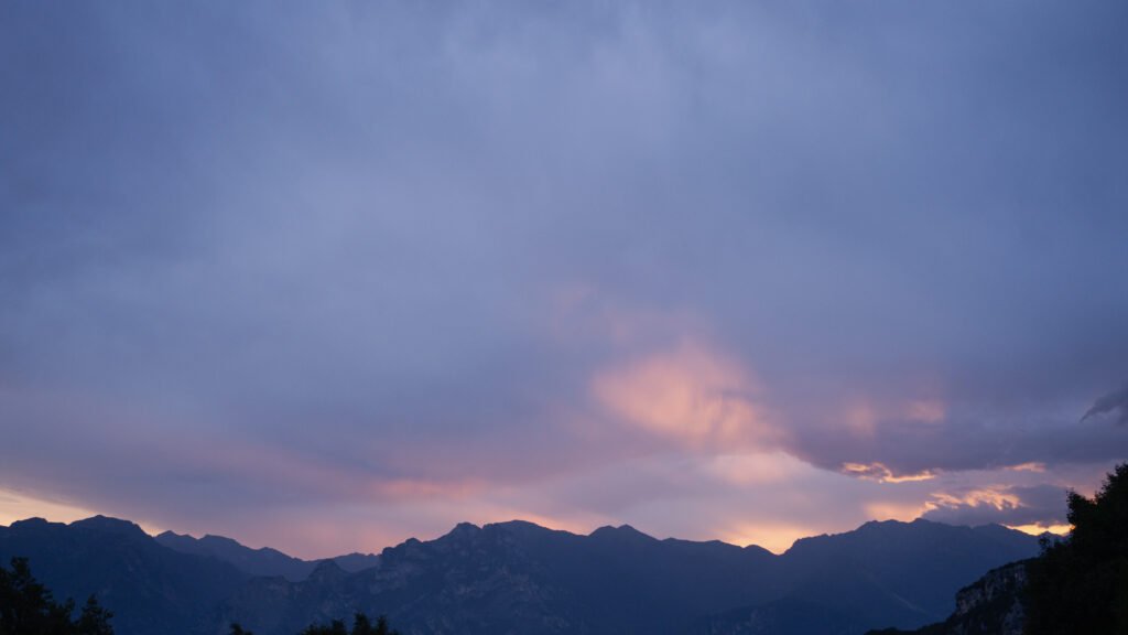 Silhouetted mountains around Lake Garda under a soft lilac dusk sky, a band of warm orange light glowing through the clouds above the ridgeline.
