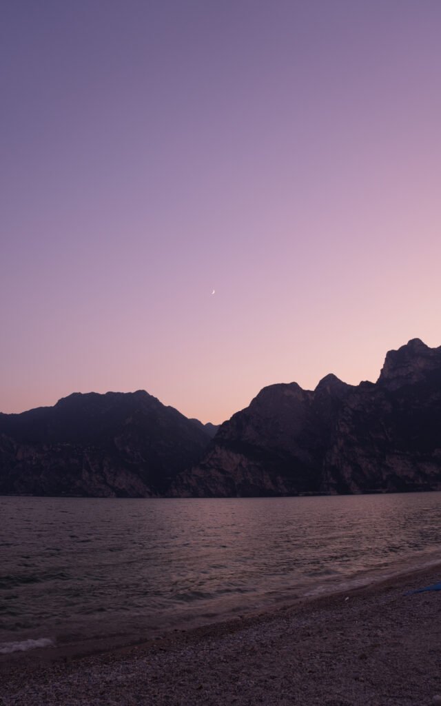 A crescent moon in a pastel pink and lilac dusk sky over the silhouetted mountains of Lake Garda, with the calm lake and a pebble beach in the foreground.
