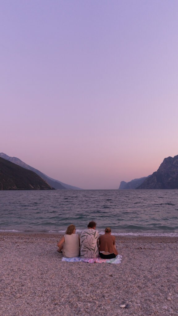 Three people sit together on a pebble beach with their backs to the camera, looking out across Lake Garda to distant mountains under a pink and lilac dusk sky.