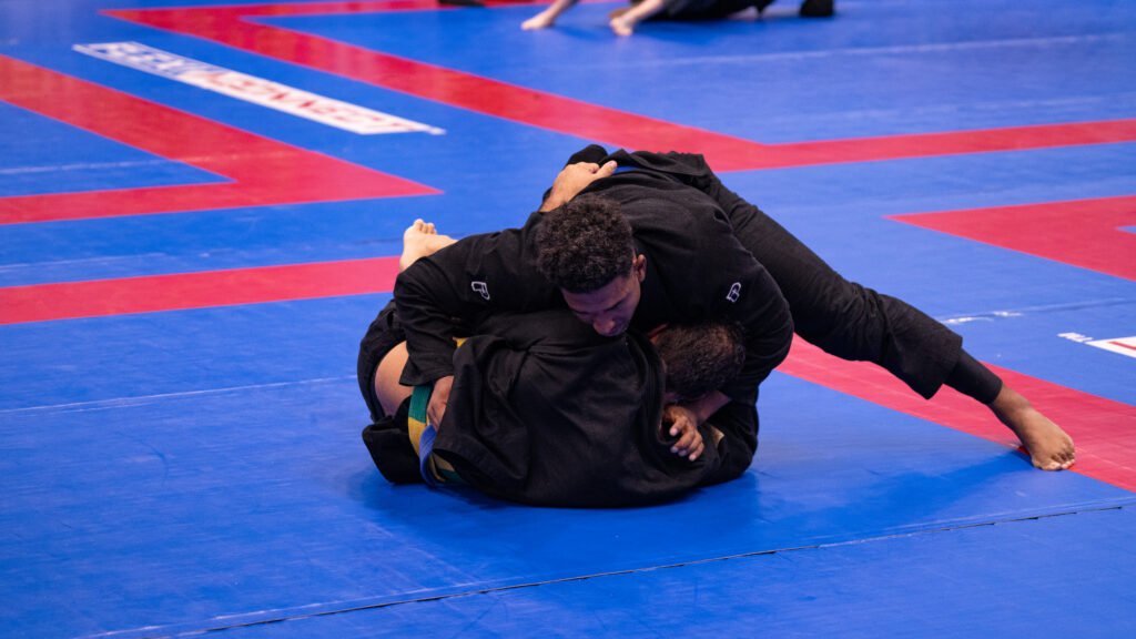 Two grapplers in black gis compete in a Brazilian jiu-jitsu match on a blue and red competition mat, one working to pass the other's guard.